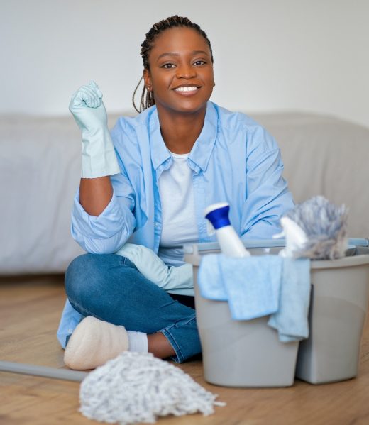 Portrait of happy pretty young black woman in homewear sitting on floor next to basket with cleaning tools in bedroom, african lady resting while doing chores at weekend at home, smiling at camera